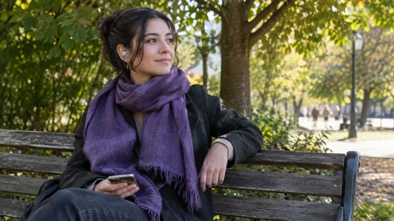 An adult woman wearing a muted purple scarf sits on a park bench beside leafy trees, listening attentively to a language partner through one small earbud.
